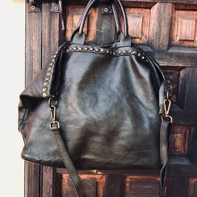 vista del bolso shopper de piel italiano con tachas marrón chocolate, colgado de una puerta de madera rústica para destacar su piel y color